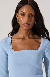 A woman is wearing a light blue, ribbed, long-sleeved top with a square neckline that has scalloped edging. She is accessorized with large, gold hoop earrings.